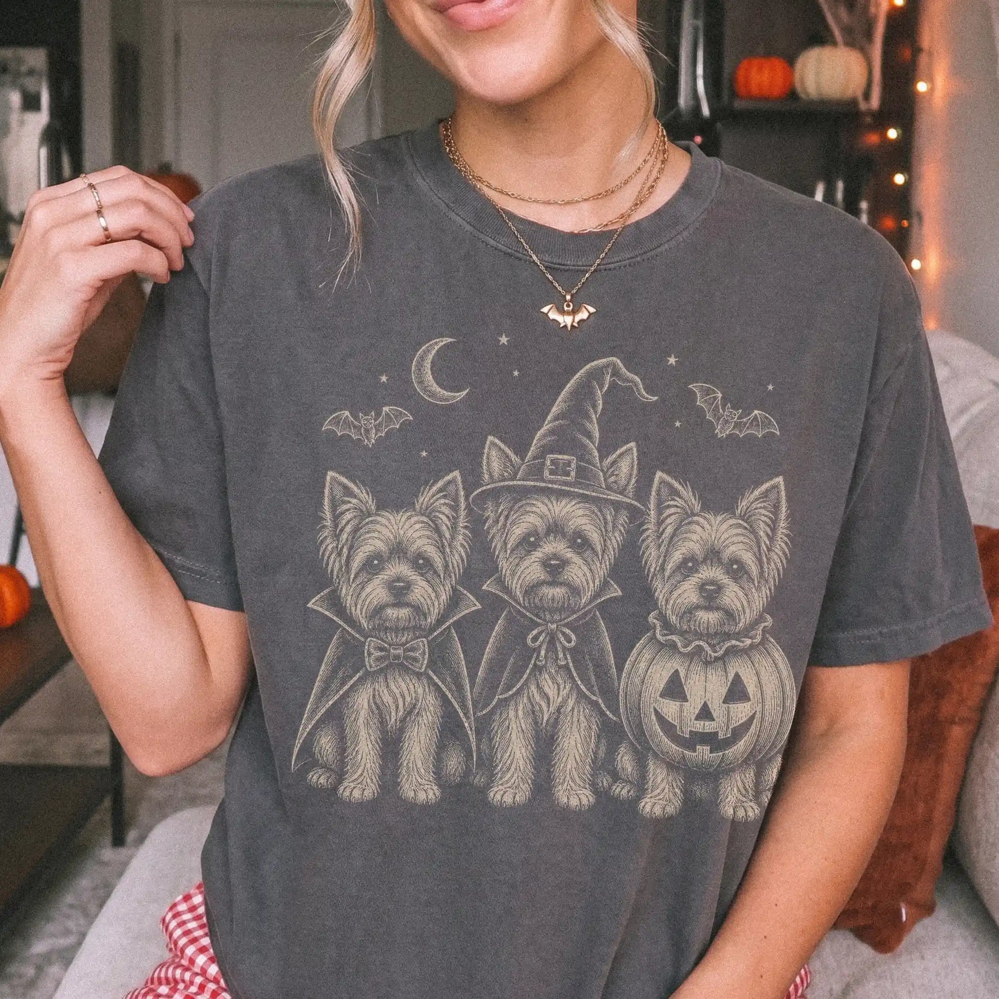 Model wearing a Pepper Comfort Colors® T-shirt showing three Yorkie dogs dressed for Halloween beneath bats and a crescent mo